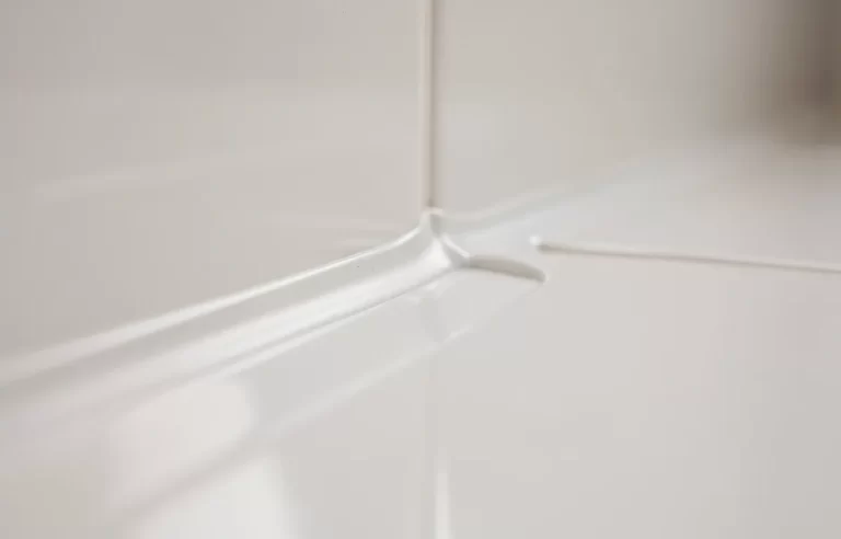 Joint en silicone soigné entre un receveur de douche moderne et des carreaux blancs brillants sous un éclairage doux.
